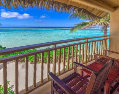 Cozy balcony with wooden chairs, overlooking a serene turquoise ocean under a clear blue sky. Palm trees frame the peaceful beach scene.