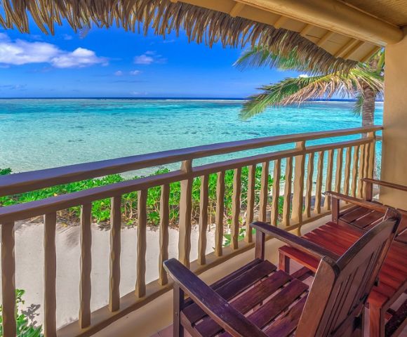 Cozy balcony with wooden chairs, overlooking a serene turquoise ocean under a clear blue sky. Palm trees frame the peaceful beach scene.