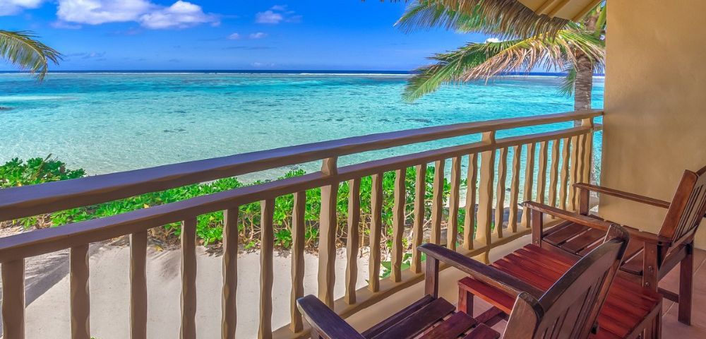 Cozy balcony with wooden chairs, overlooking a serene turquoise ocean under a clear blue sky. Palm trees frame the peaceful beach scene.