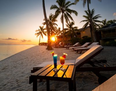 Sunset beach scene with palm trees and lounge chairs on white sand. Two colorful cocktails sit on a table, creating a relaxed, tropical vibe.