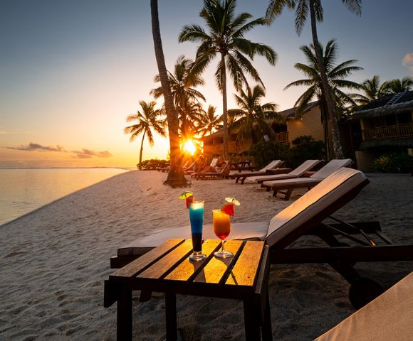 Sunset beach scene with palm trees and lounge chairs on white sand. Two colorful cocktails sit on a table, creating a relaxed, tropical vibe.