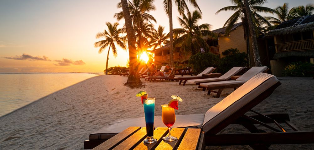Sunset beach scene with palm trees and lounge chairs on white sand. Two colorful cocktails sit on a table, creating a relaxed, tropical vibe.