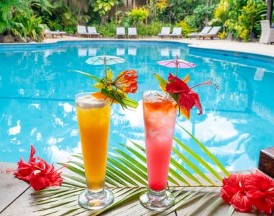 Two tropical cocktails, orange and pink, with decorative umbrellas and hibiscus flowers, sit by a clear blue pool, surrounded by lush greenery.