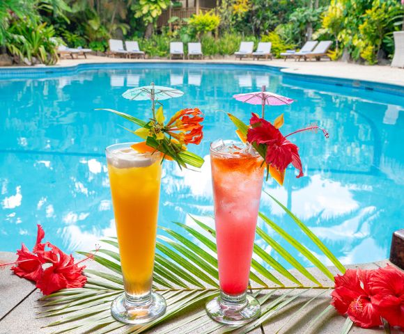 Two tropical cocktails, orange and pink, with decorative umbrellas and hibiscus flowers, sit by a clear blue pool, surrounded by lush greenery.