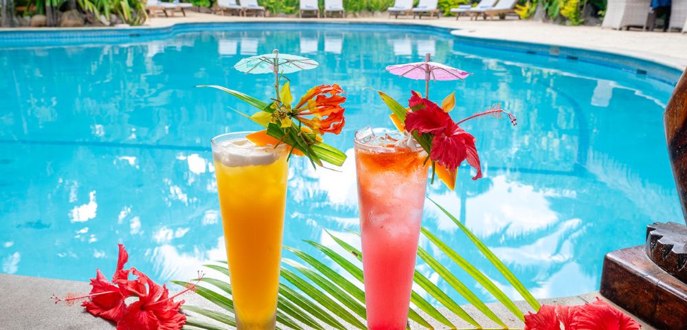 Two tropical cocktails, orange and pink, with decorative umbrellas and hibiscus flowers, sit by a clear blue pool, surrounded by lush greenery.