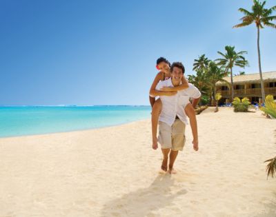 A joyful couple enjoys a sunny beach day; one gives the other a piggyback ride on a sandy shore. Clear blue sky, turquoise sea, and palm trees enhance the tropical vibe.