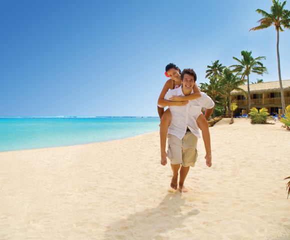 A joyful couple enjoys a sunny beach day; one gives the other a piggyback ride on a sandy shore. Clear blue sky, turquoise sea, and palm trees enhance the tropical vibe.