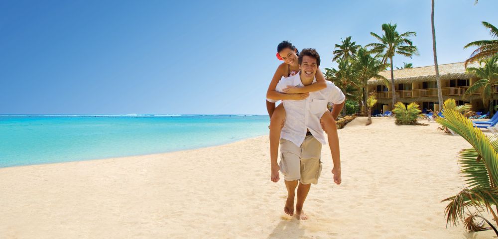 A joyful couple enjoys a sunny beach day; one gives the other a piggyback ride on a sandy shore. Clear blue sky, turquoise sea, and palm trees enhance the tropical vibe.