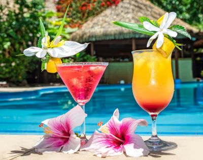 Two tropical cocktails by a poolside. A pink drink and an orange drink, each garnished with flowers, create a vibrant and refreshing summer vibe.