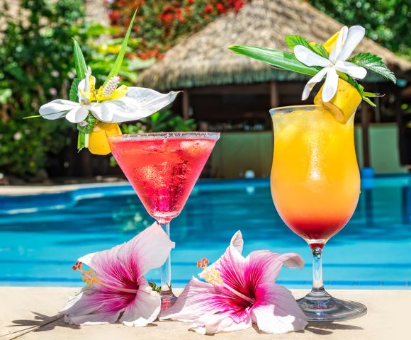 Two tropical cocktails by a poolside. A pink drink and an orange drink, each garnished with flowers, create a vibrant and refreshing summer vibe.
