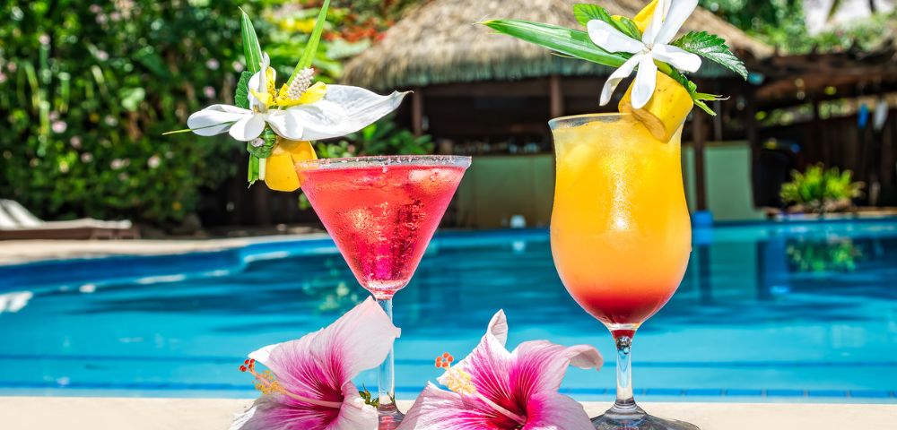 Two tropical cocktails by a poolside. A pink drink and an orange drink, each garnished with flowers, create a vibrant and refreshing summer vibe.