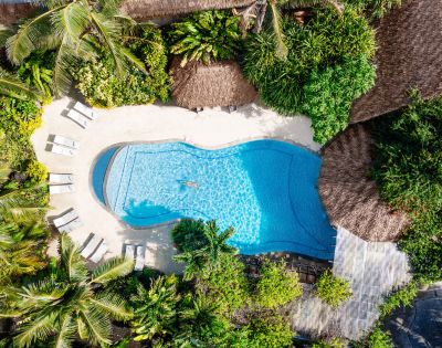 Aerial view of a serene pool surrounded by lush greenery and thatched roofs. Lounge chairs are placed around the pool, creating a relaxing tropical oasis.