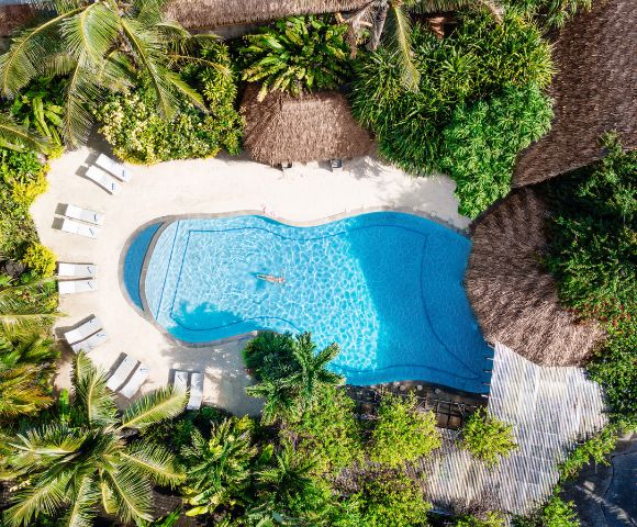 Aerial view of a serene pool surrounded by lush greenery and thatched roofs. Lounge chairs are placed around the pool, creating a relaxing tropical oasis.