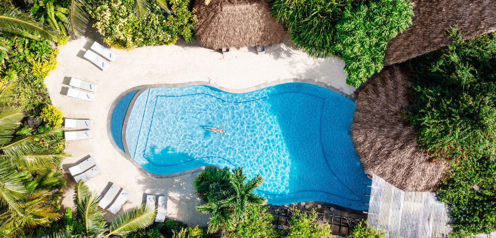 Aerial view of a serene pool surrounded by lush greenery and thatched roofs. Lounge chairs are placed around the pool, creating a relaxing tropical oasis.