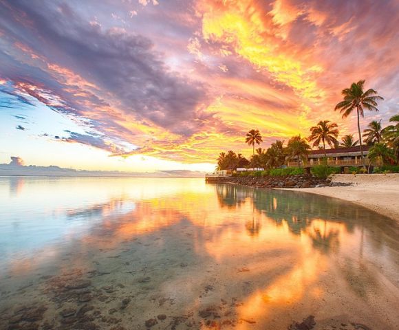 Sunset over a tropical beach with vibrant orange and pink skies reflecting on calm water, surrounded by palm trees and beachfront houses.