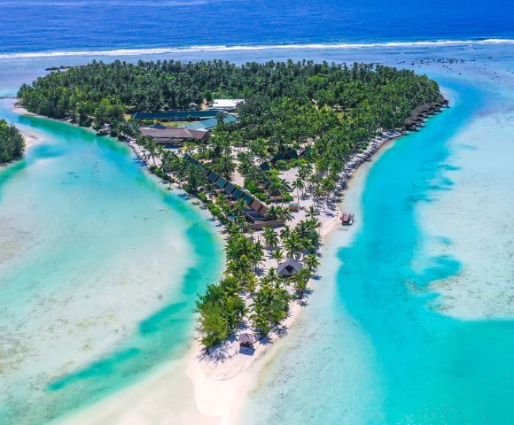 Aerial view of a tropical island with lush greenery, bordered by turquoise water and sandy beaches. Numerous overwater bungalows add to the serene, luxurious feel.
