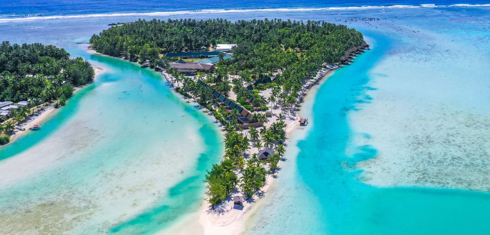 Aerial view of a tropical island with lush greenery, bordered by turquoise water and sandy beaches. Numerous overwater bungalows add to the serene, luxurious feel.