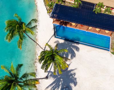 Aerial view of a tropical beach with turquoise water, palm trees, and a rectangular swimming pool beside a sunlit deck. Relaxed, vacation vibe.