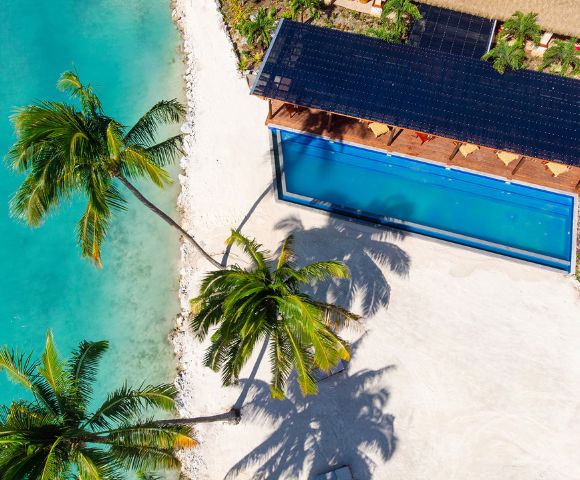 Aerial view of a tropical beach with turquoise water, palm trees, and a rectangular swimming pool beside a sunlit deck. Relaxed, vacation vibe.
