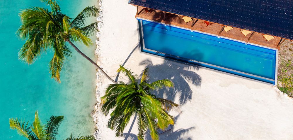 Aerial view of a tropical beach with turquoise water, palm trees, and a rectangular swimming pool beside a sunlit deck. Relaxed, vacation vibe.