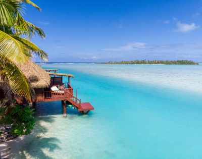 Thatched-roof hut on stilts over clear turquoise water, surrounded by lush palm trees and distant islands, under a bright blue sky, conveying tranquility.