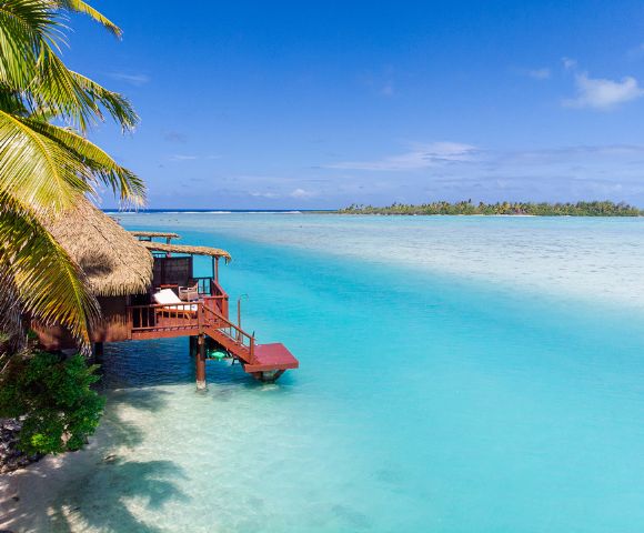 Thatched-roof hut on stilts over clear turquoise water, surrounded by lush palm trees and distant islands, under a bright blue sky, conveying tranquility.