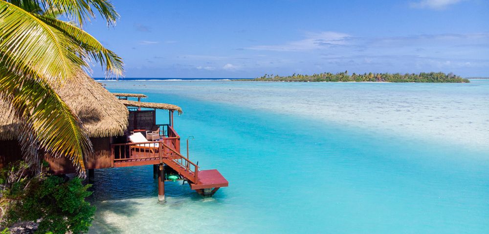 Thatched-roof hut on stilts over clear turquoise water, surrounded by lush palm trees and distant islands, under a bright blue sky, conveying tranquility.