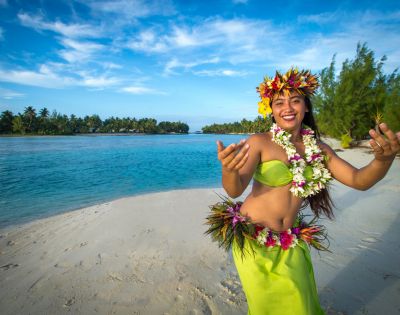 A smiling woman in traditional Hawaiian attire dances on a beach, with turquoise water and lush greenery under a bright blue sky in the background.
