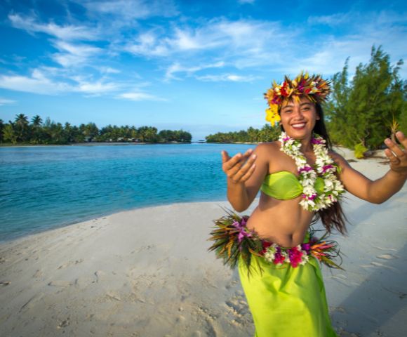 A smiling woman in traditional Hawaiian attire dances on a beach, with turquoise water and lush greenery under a bright blue sky in the background.