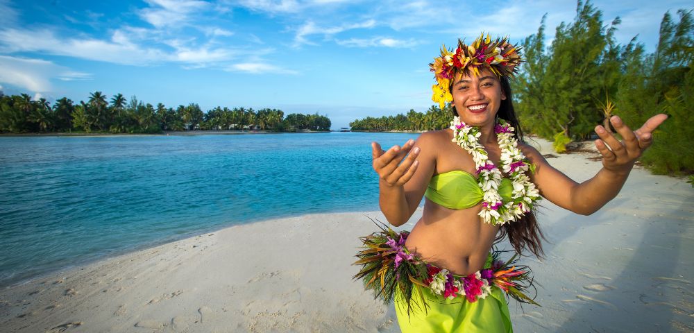 A smiling woman in traditional Hawaiian attire dances on a beach, with turquoise water and lush greenery under a bright blue sky in the background.