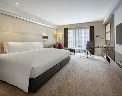 Modern hotel room with a large bed, crisp white linens, and a stylish workspace. Natural light streams through floor-to-ceiling windows, creating a bright, inviting atmosphere.