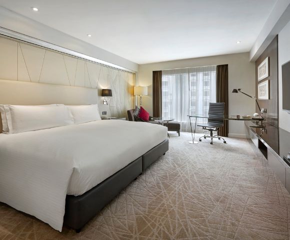 Modern hotel room with a large bed, crisp white linens, and a stylish workspace. Natural light streams through floor-to-ceiling windows, creating a bright, inviting atmosphere.