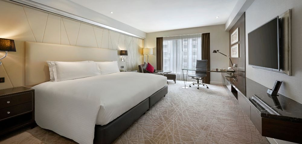 Modern hotel room with a large bed, crisp white linens, and a stylish workspace. Natural light streams through floor-to-ceiling windows, creating a bright, inviting atmosphere.
