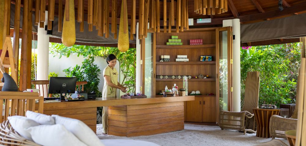 A woman in uniform arranges items on a wooden reception desk in a tranquil spa. Soft natural light, modern decor, and lush greenery create a calming atmosphere.