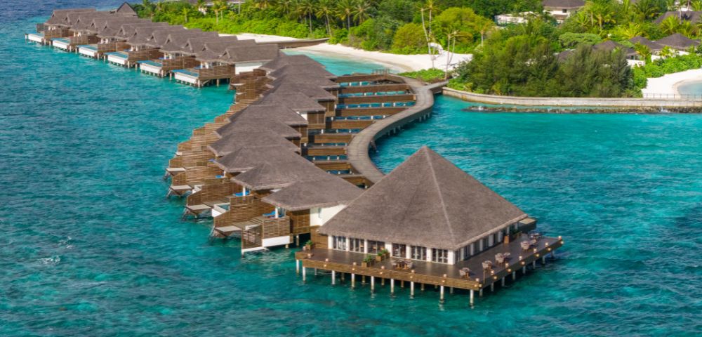 Overwater bungalows with thatched roofs stretch in a curve over turquoise waters, surrounded by lush greenery, evoking a serene, tropical ambiance.