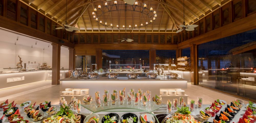 Luxurious buffet in a spacious, warmly lit dining hall. Elegant food displays on glass tables, high wooden ceiling, and ambient, inviting atmosphere.