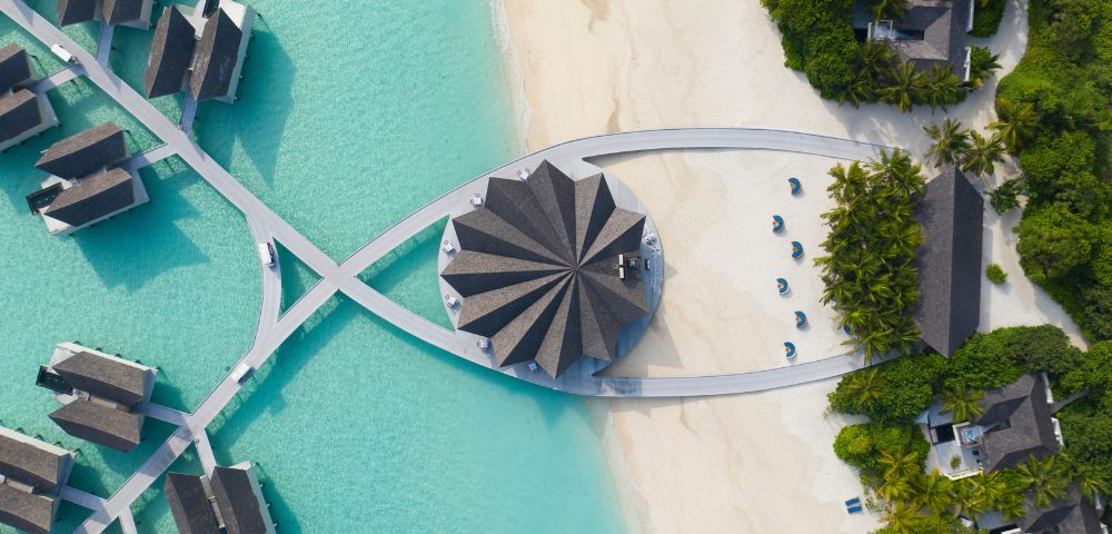 Aerial view of a tropical resort with overwater villas on turquoise water, a central pavilion with a star-shaped roof, and a sandy beach with lounge chairs.