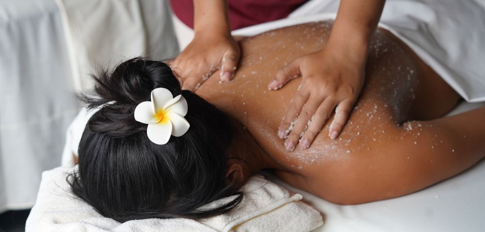 A person receives a back massage with exfoliating scrub, adorned with a frangipani flower in their hair. The scene conveys relaxation and spa serenity.