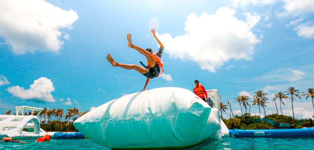 A person in an orange life jacket is launched into the air from a giant inflatable bag over water. A clear blue sky and palm trees are in the background.