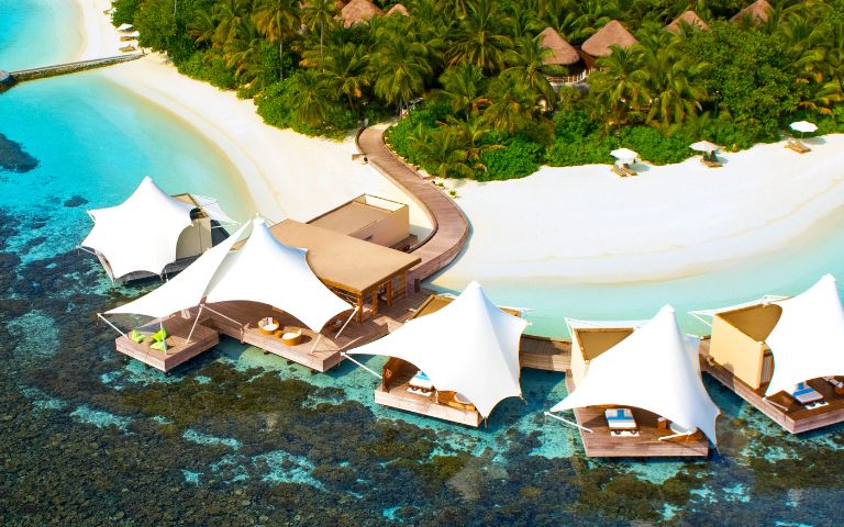 Aerial view of a tropical island resort with white sand beach, lush greenery, and turquoise waters. Luxurious overwater cabanas line the shore, creating a serene, idyllic setting.