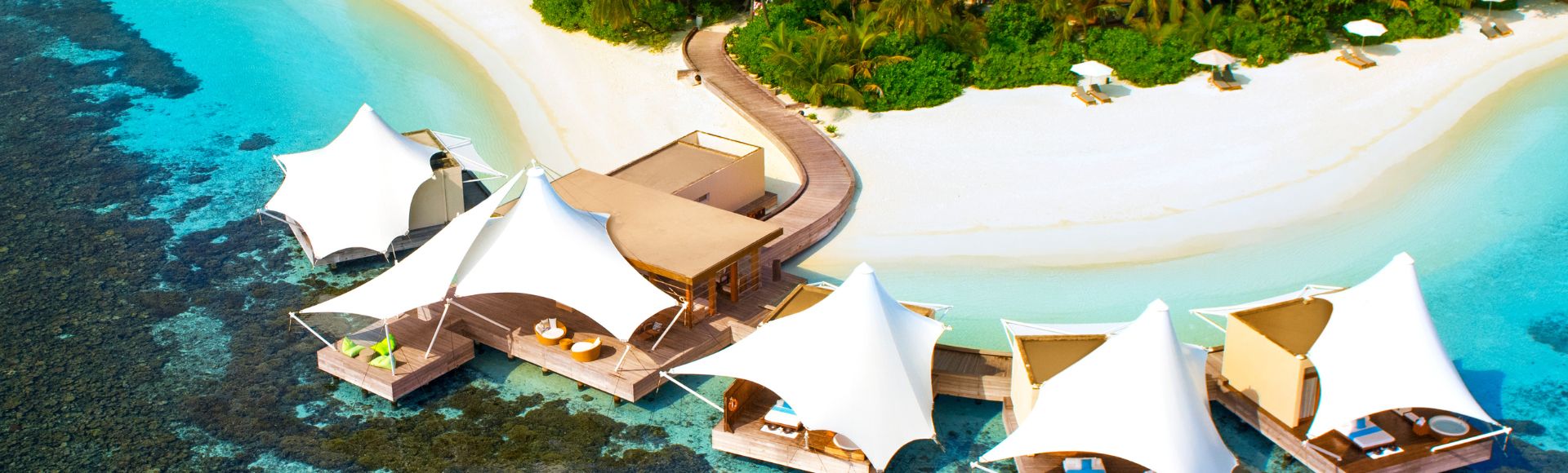 Aerial view of a tropical island resort with white sand beach, lush greenery, and turquoise waters. Luxurious overwater cabanas line the shore, creating a serene, idyllic setting.