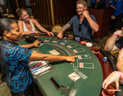 A lively casino scene with smiling people playing blackjack. Dealer manages the cards, while players enjoy drinks and engage in the game.