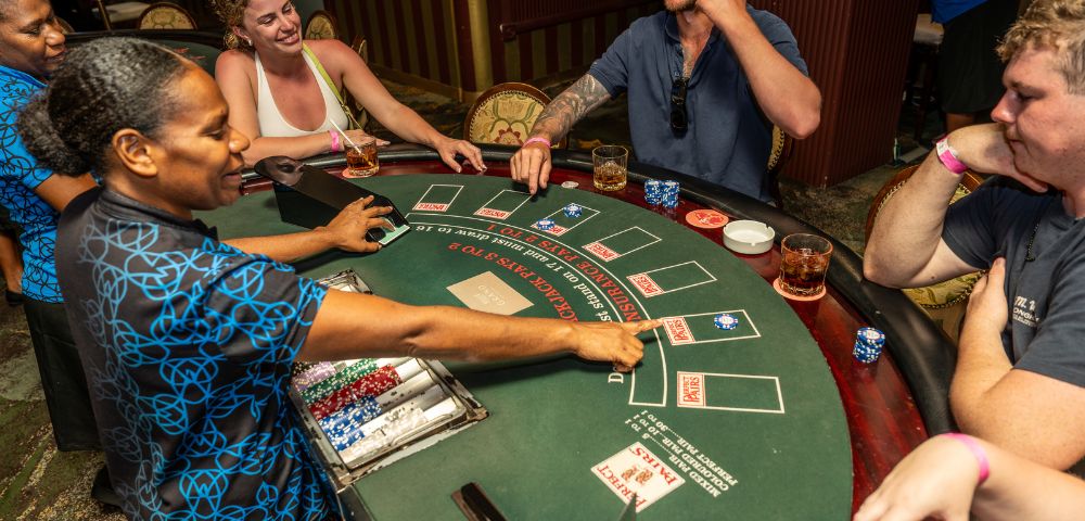 A lively casino scene with smiling people playing blackjack. Dealer manages the cards, while players enjoy drinks and engage in the game.