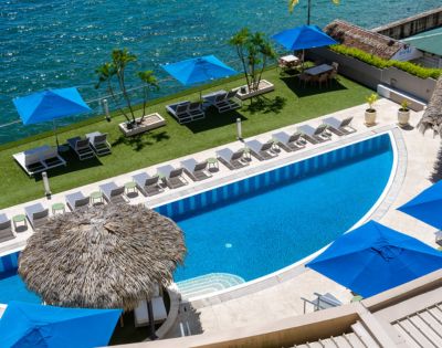 A serene poolside scene with a curved swimming pool overlooking a blue ocean. Sun loungers, umbrellas, and palm trees create a tropical atmosphere.