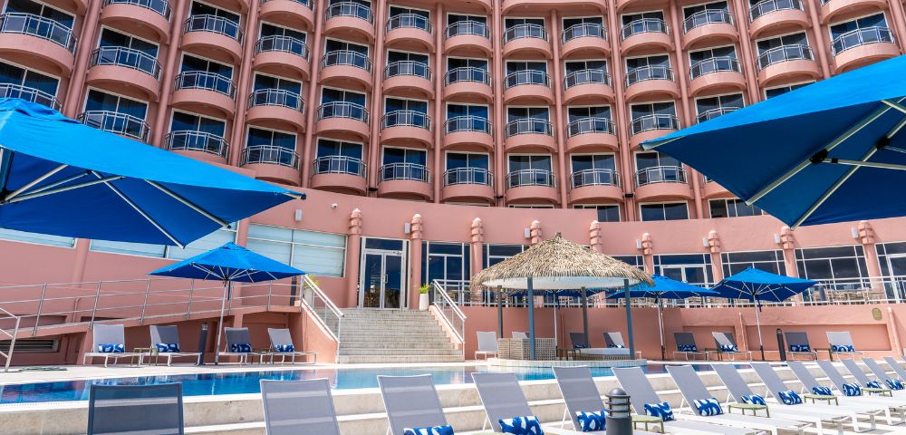 A pink hotel with curved balconies rises above a pool deck, featuring blue lounge chairs and umbrellas. The scene is sunny and inviting.