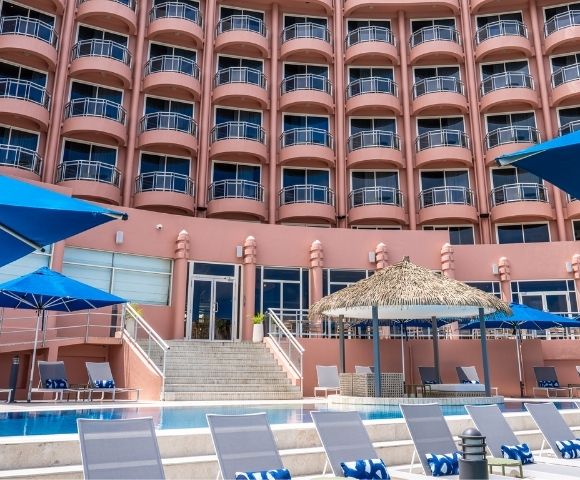 Large hotel with multiple balconies, set under a partly cloudy sky. In the foreground, blue umbrellas and poolside lounge chairs create a relaxed atmosphere.