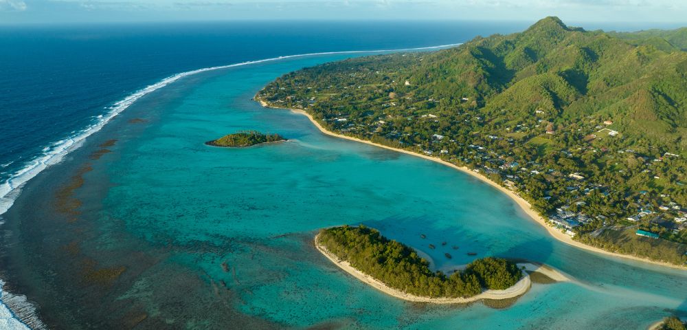 Aerial view of a tropical coastline with vibrant turquoise waters, lush green mountains, and small islands. The scene is peaceful and picturesque.