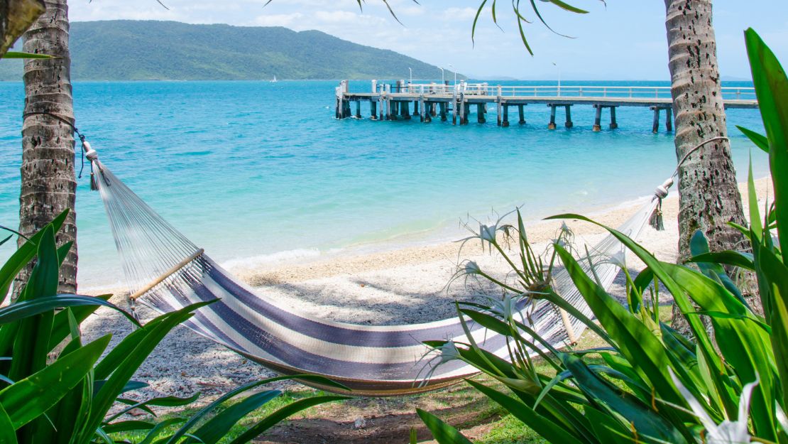 A serene beach scene featuring a striped hammock between palm trees, overlooking turquoise waters and a wooden pier. Lush greenery frames the peaceful view.