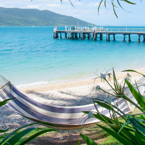 A serene beach scene featuring a striped hammock between palm trees, overlooking turquoise waters and a wooden pier. Lush greenery frames the peaceful view.