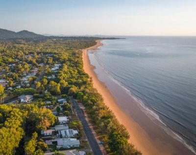 Aerial view of a serene coastline during sunset. The golden sandy beach curves alongside calm blue waters, bordered by lush green trees and a small coastal town.